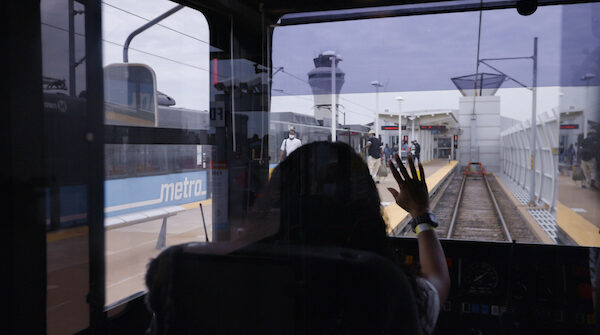 Interior shot of a MetroLink operator waving to riders on the platform of the Lambert Airport Terminal #1 MetroLink station. The platform and airport control tower can be seen through the windshield.